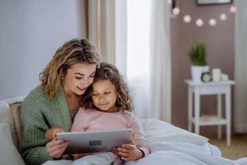 Happy mother with her little daughter lying on bed and using tablet at home.