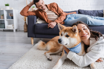 Wall Mural - cheerful young woman hugging akita inu dog near boyfriend on couch.