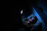 Fototapeta Zwierzęta - Pretty, middle-aged woman using her cell phone in bed at night - unhealthy blue light exposure