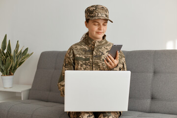 Wall Mural - Horizontal shot of Caucaian military woman wearing camouflage uniform and cap sitting on sofa and working on laptop, holding using smartphone, looking at device screen.