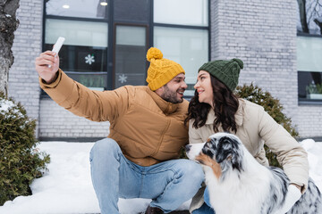 Wall Mural - happy man in knitted hat taking selfie with cheerful girlfriend and australian shepherd dog in winter.