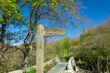 Public footpath sign to Satron, a rural hamlet in Swaledale, with 16th Century Ivelet Bridge in the background.   Springtime in the Yorkshire Dales with blue sky and trees bursting into leaf. 
