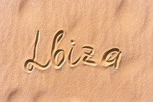 The Text "Ibiza" In The Center Of The Photo Is Handwritten On A Golden Sandy Beach Near The Sea. Next To The Text Are Waves Of Sand. Flat Lay Photo Texture.