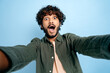 © Kateryna - Joyful positive Indian funny guy with gladden face expression, in casual shirt, doing selfie shot on smartphone and fooling around into the camera, stands on isolated blue background