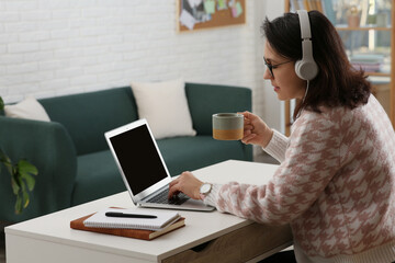 Poster - Woman with modern laptop and headphones drinking tea while learning at table indoors