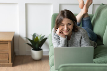 Wall Mural - Young beautiful smiling woman using laptop lying on the couch. Concept of freelance job, home office. Female using technology for learning, shopping, e-commerce, communication, social media