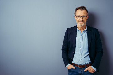 smiling bestager with glasses stands in front of a blue wall and looks into the camera