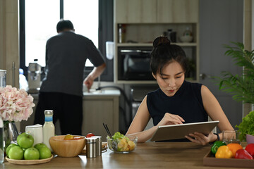 Wall Mural - Smiling millennial woman sitting at table at home kitchen and using digital tablet.