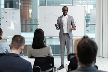 Poster - Young African American male coach explaining financial data to audience while standing by whiteboard with graphs in lecture hall