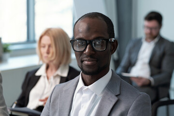 Poster - Young contemporary black man in formalwear and eyeglasses looking at camera while sitting against male and female colleagues