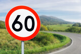 Fototapeta  - Speed limit sign with curvy road behind. Maximum sixty kilometers per hour. Safety on road background. White round sign red border line. Traffic ticket background. Speeding fine. Slow down on turn.
