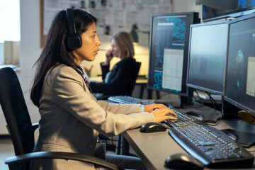 Side view of young Hispanic female operator of surveillance system sitting by desk in front of computer monitors in office