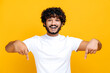 © Kateryna - Amazed indian or arabian cheerful young man in white basic t-shirt looks at the camera and points fingers down at space for your presentation, stands on isolated orange color background, smiling