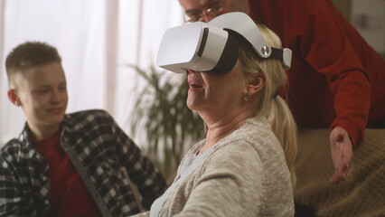 Cheerful grandmother in virtual reality glasses on sofa laughing and gesturing near happy grandson and grandfather in living room