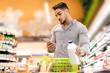 © Prostock-studio - Arabic Guy Using Cellphone Holding Milk Buying Groceries In Supermarket