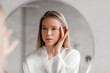 © Prostock-studio - Pretty young woman looking at mirror and touching face while doing morning beauty routine in bathroom