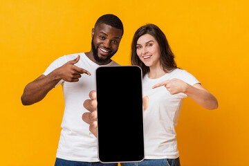 Use this new application. Happy multiracial couple pointing at smartphone with blank screen, mockup for design