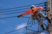 Low Angle View Of Electrician With Safety Equipment And Various Work Tools Is Installing Cable Lines And Electrical System On Electric Power Pole Against Blue Sky Background