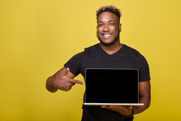 Satisfied African holds modern laptop in his hands and points his finger black screen where you can leave your ads. Dark-skinned man smiles and draws his hand attention to laptop he holds in his hand