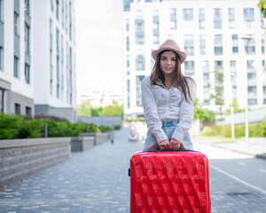 Beautiful caucasian brunette woman in a hat posing with a suitcase on a city street.