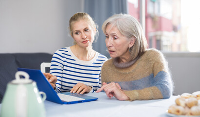 Mature woman and her adult daughter, sitting at a table, are viewing the necessary information on a laptop