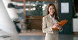 © NAMPIX - Smiling young businesswoman looking at camera while standing in the coworking space.