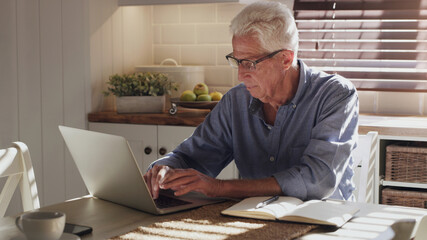 I better get this to the client quick. Shot of a senior man sitting alone in the kitchen and using his laptop to work from home.