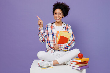 Wall Mural - Full body young girl woman of African American ethnicity student in shirt sit hold books point finger aside workspace area isolated on plain purple background Education in university college concept.