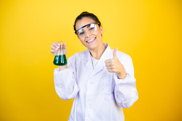 Young brunette woman wearing scientist uniform holding test tube over isolated yellow background success sign doing positive gesture with hand, thumb up smiling and happy. cheerful expression