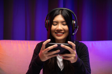 Playing video game on cellphone. Young asian pretty woman sitting on sofa holding smartphone in living room. Happy female Pro Streamer chinese wearing headphone playing game online with neon light.