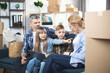 © sofiko14 - Happy big Caucasian family planning design on new apartment. Parents with their two kids sitting together on couch and using modern laptop. Moving boxes around. Cute girl pointing on the box