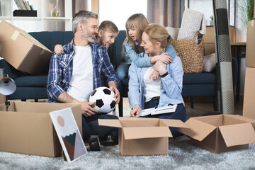 Wall Mural - Mature Caucasian parents with their two teen kids, boy and girl, posing among boxes at new apartment. Happy big family, hugging and looking each other, enjoying mowing day.