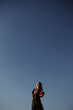 © Alex - Against the background of the sky poses a Caucasian girl wearing a black dress, overall plan, front view.