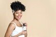 © etonastenka - Portrait of a beautiful black woman holding a bottle of liquid foundation on a beige background. African american young woman smiling while holding bottle of foundation