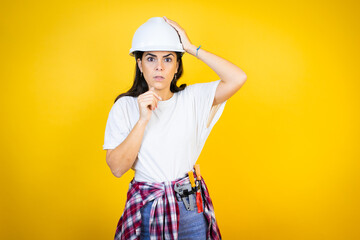 Wall Mural - Young caucasian woman wearing hardhat and builder clothes over isolated yellow background putting one hand on her head smiling like she had forgotten something