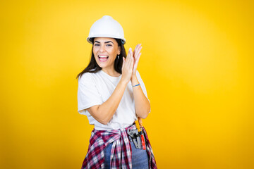 Young caucasian woman wearing hardhat and builder clothes over isolated yellow background clapping and applauding happy and joyful, smiling proud hands together