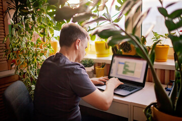 a man working with laptop remotely from home. a distant work place with many home plants. green natu