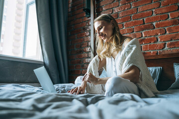 Adult woman forty years freelancer with blonde long hair in casual clothes using laptop sitting on bed at the home