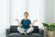 © PaeGAG - Happy calm Asian woman sit on sofa at home meditation with mudra hands in lotus pose. Smiling young Asian woman relax on couch in living room practice yoga, relieve negative emotions.