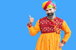 Happy beard Indian man wear traditional colorful outfit and turban pointing at copy space, standing against blue background. rajasthan male customer advertising shopping sale, discount, promo.