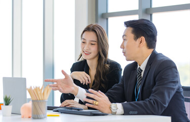 Asian young happy professional successful businessman businesswoman employee partnership in formal business suit sitting at working desk in company meeting room smiling discussing talking together