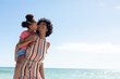 Leinwandbild Motiv African american girl kissing mother while enjoying piggyback ride on her at beach against blue sky