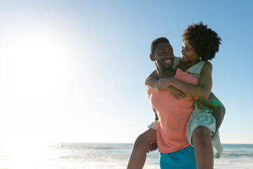 African american man giving piggyback to girlfriend at beach against sky with copy space