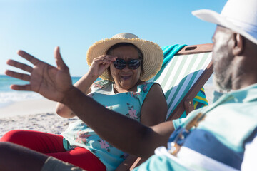 Wall Mural - Retired african american senior couple sitting on folding chairs talking at beach during sunny day