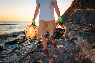 Wall Mural - Close up of volunteer walking along at the wild beach holding two plastic bags. In the background, the ocean and the sunset. The concept of cleaning the beach from pollution