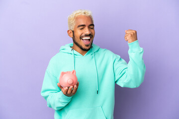 Canvas Print - Young Colombian man holding a piggybank isolated on purple background celebrating a victory