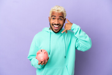 Wall Mural - Young Colombian man holding a piggybank isolated on purple background making phone gesture. Call me back sign