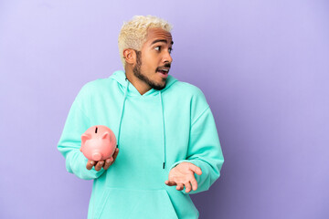 Canvas Print - Young Colombian man holding a piggybank isolated on purple background with surprise expression while looking side