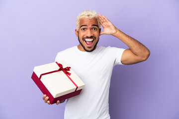 Sticker - Young Colombian man holding a gift isolated on purple background with surprise expression