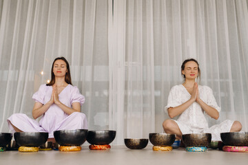 Two women are sitting with Tibetan bowls in the lotus position before a yoga class in the gym